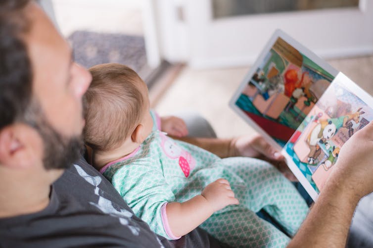 Un bébé vêtu d'une dormeuse vert menthe est assis sur les genoux de son père pendant qu'il lui lit un livre.