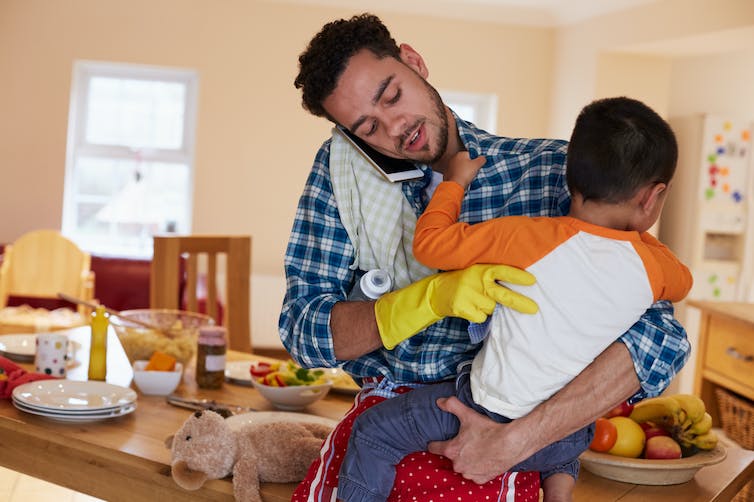 Un homme parle dans son téléphone portable tout en tenant un enfant dans ses bras.  Il a un gant de nettoyage dans une main et un biberon sous le bras.