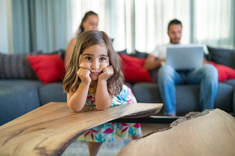 Mère et père utilisant un ordinateur portable sur un canapé avec une fille qui s'ennuie au premier plan.