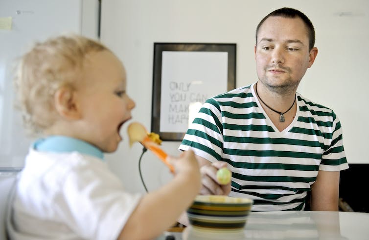 Un homme en T-shirt rayé regarde son fils manger dans sa chaise haute.