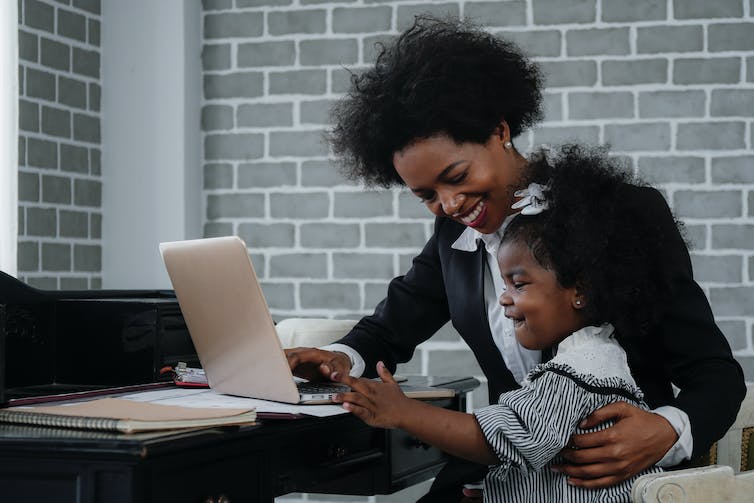 Un enfant est assis sur les genoux d'une femme, vraisemblablement sa mère, devant un bureau. Elle sourit et touche un ordinateur portable tandis que sa mère lui sourit.