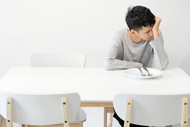 Un adolescent assis à une table blanche avec une assiette blanche vide dessus, posant sa tête sur sa main