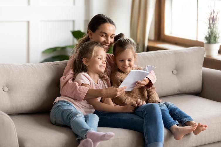 Une jeune femme assise sur un canapé avec deux filles lisant un livre.