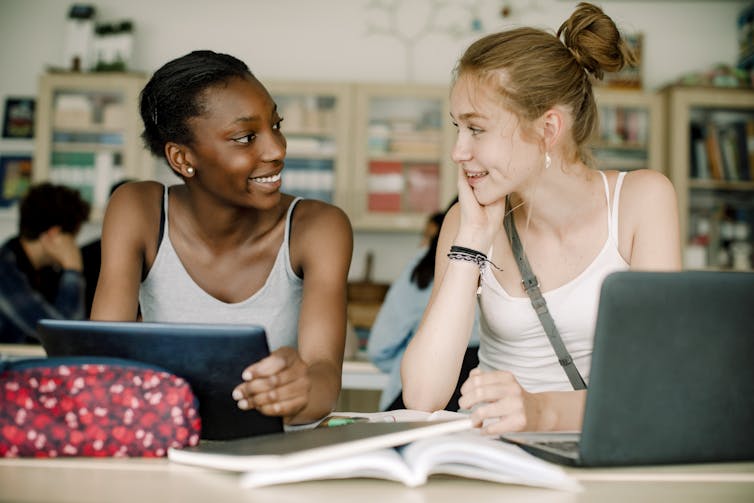 Deux adolescentes en débardeur blanc sourient et discutent alors qu'elles sont assises à une table dans une salle de classe.