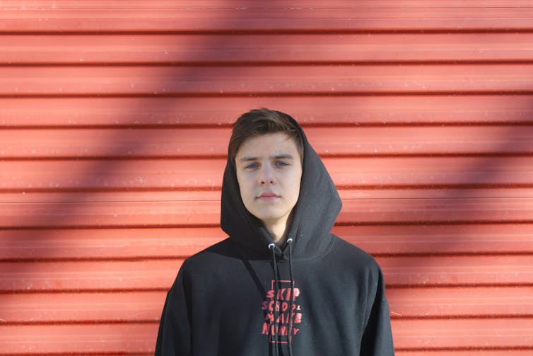 Un jeune homme se tient debout contre un mur en bois rouge. Il porte un pull à capuche noir.