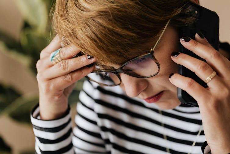 Une femme porte un téléphone portable à son oreille. Elle a une expression sérieuse et une main sur la tempe.