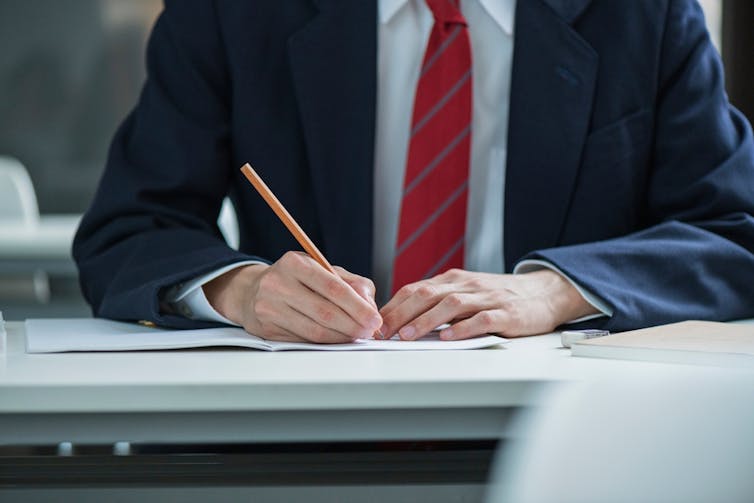 Un adolescent en blazer et cravate écrit sur une feuille de papier posée sur un bureau.