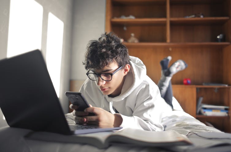 Un adolescent est allongé sur un lit avec un ordinateur portable et un livre, tout en effectuant un test sur un téléphone.