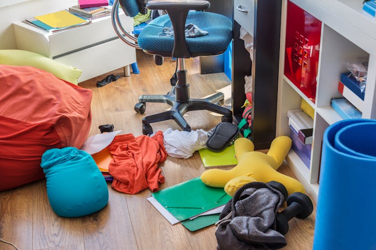 Des vêtements, des papiers et des livres sur le sol, près d'une chaise de bureau et d'étagères.