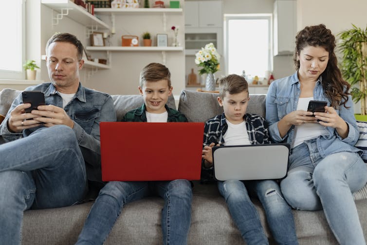 un homme et une femme sur des smartphones sont assis sur un canapé avec deux enfants utilisant des ordinateurs portables, tous absorbés par les écrans
