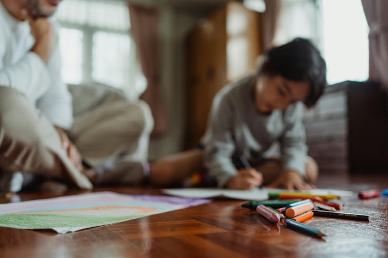 Un jeune enfant et un homme sont assis par terre avec des crayons de couleur et des papiers. L'enfant dessine sous le regard de l'homme.