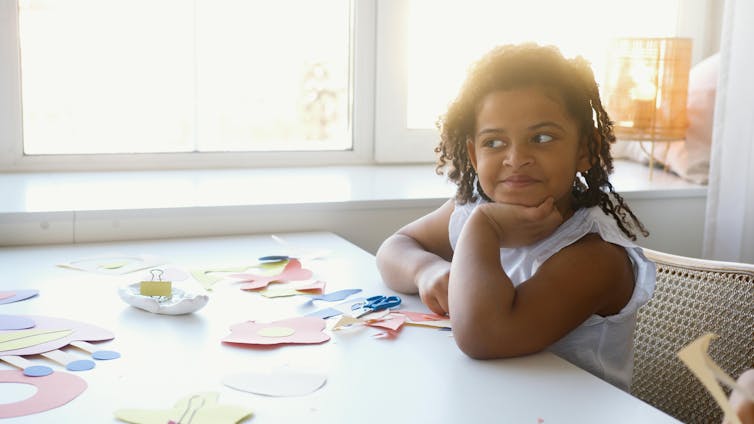 Une jeune fille pose son menton dans sa main. Il y a des ciseaux et des papiers de couleur sur une table.