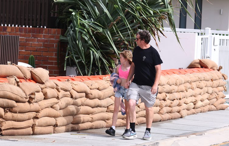 Un homme et une jeune fille passent devant une rangée de sacs de sable dans une rue de banlieue.