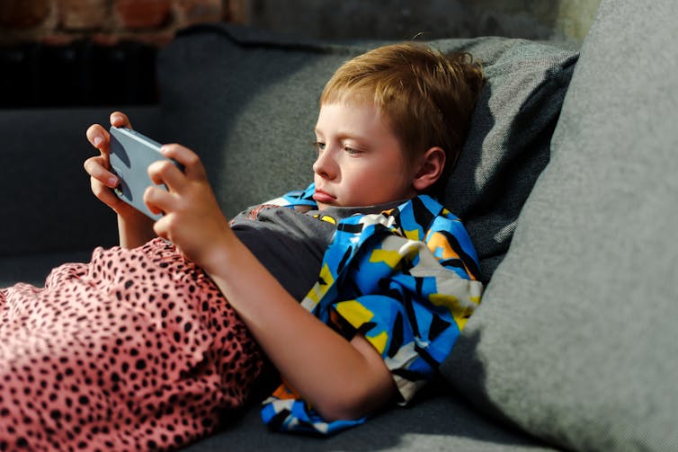 Un jeune garçon fait la moue, tandis de coucher sur le canapé avec un téléphone et une couverture.