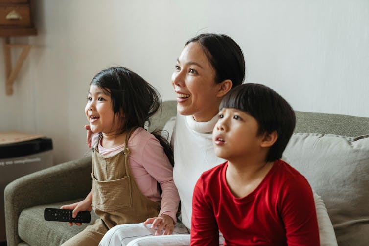 Une femme et deux jeunes enfants regardent la télévision sur le canapé.