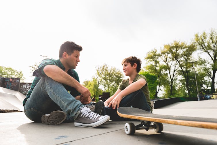 Un homme adulte et un enfant s'asseyent et parlent dans un skate park.