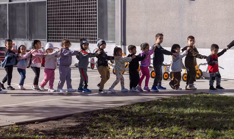 Un groupe de jeunes enfants marchent en ligne, les mains sur les épaules de l’enfant devant eux.