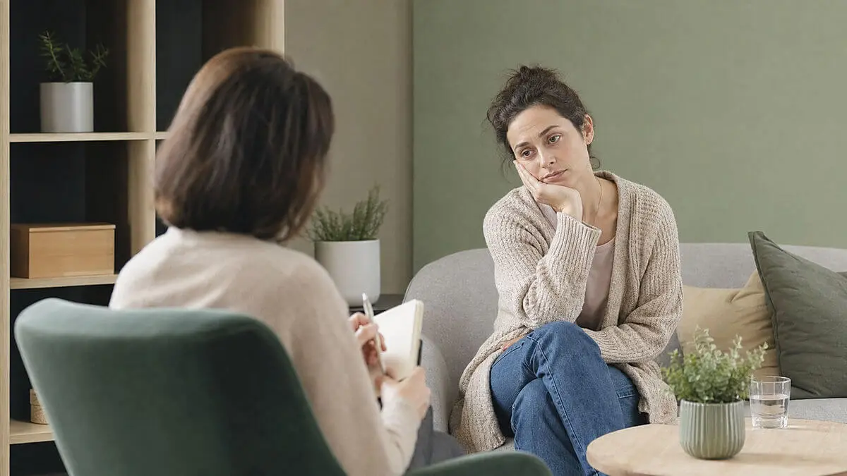 Intérieur d'une consultation calme, thérapeute assis devant une mère fatiguée avec un carnet, geste empathique, composition moyenne, lumière douce
