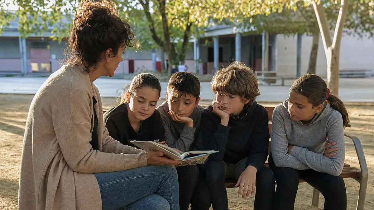 Cour de récréation au coucher du soleil : petit groupe d'enfants de 9 ans lisant une histoire assis sur un banc, un enseignant guide avec geste, composition